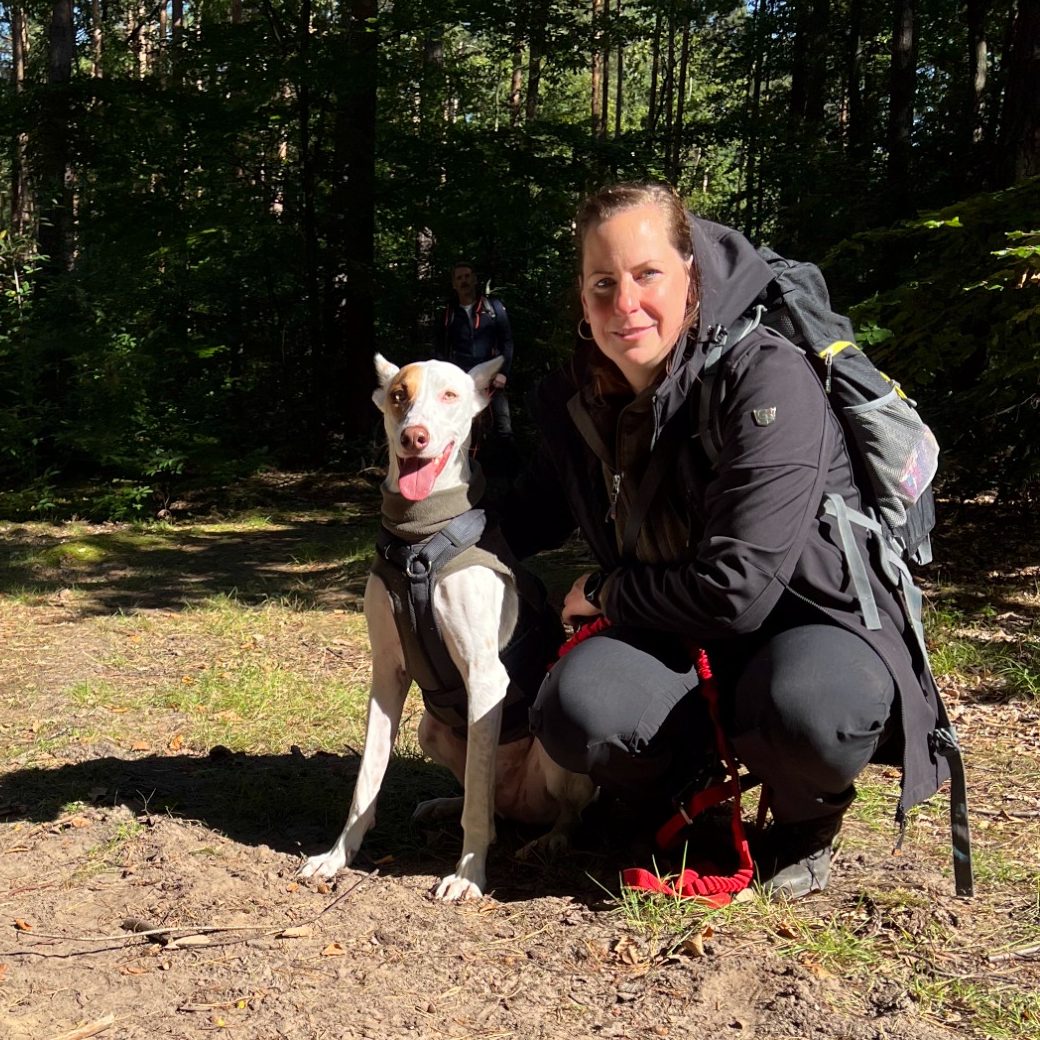 Frau mit Rucksack kniet neben einem Hund in einem Wald. Sonne scheint durch Bäume.